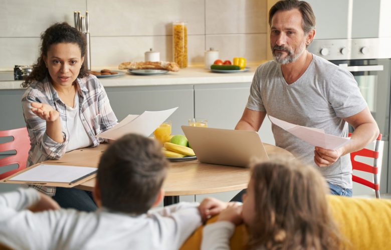 Hispanic mother and Caucasian father at table with documents speaking to two kids on sofa